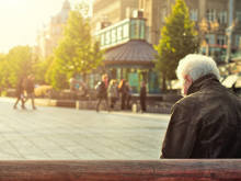 Älterer Heer sitzt mit dem Rücken zur Kamera auf eine Bank, vor ihm ein Stadtplatz auf dem gerade die Sonne aufgeht