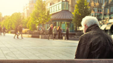 Älterer Heer sitzt mit dem Rücken zur Kamera auf eine Bank, vor ihm ein Stadtplatz auf dem gerade die Sonne aufgeht