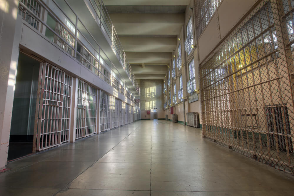 Langer Gang in einem Gefängnis, rechts und links befinden sich teils offene Zellentüren.