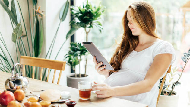 Eine schwangere Frau frühstückt am Tisch und schaut in ein Tablet.