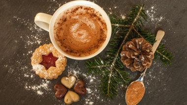 Kakao in einer weißen Kaffeetasse mit Weihnachtsplätzchen und einem Tannenzweig auf einem Holzuntergrund.