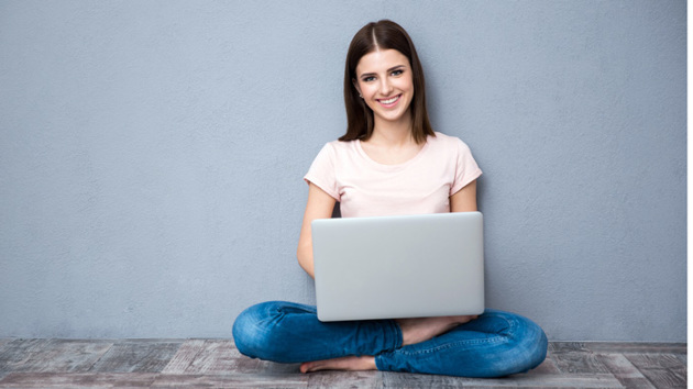 Eine junge Frau sitzt mit ihrem Laptop auf dem Boden vor einer grauen Wand und lächelt.