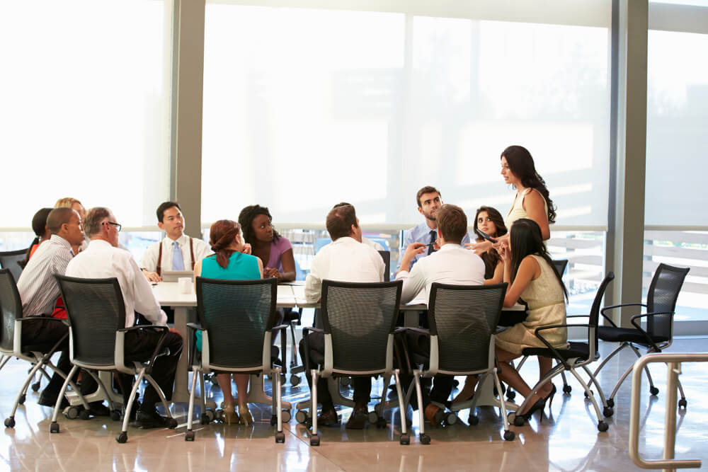 Eine Gruppe von Mitarbeitern einer Firma hat sich um einen großen Konferenztisch versammelt und blickt gebannt auf die Vorgesetzte, die am Kopf des Tisches steht.