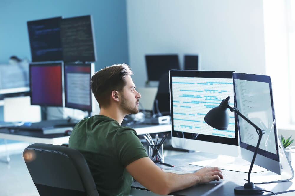 Ein Programmierer sitzt an seinem Arbeitsplatz vor mehreren Bildschirmen und erstellt eine Software.