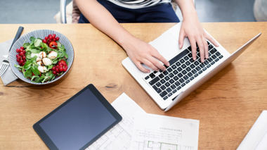 Frau am Schreibtisch mit Laptop neben einem Bauplan und Salat