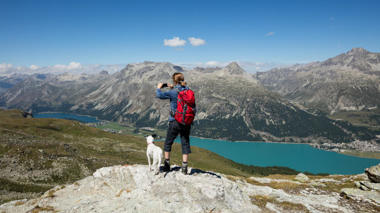 Eine Frau steht mit einem Hund auf einem Berg und macht mit ihrem Smartphone ein Bild von der Landschaft.