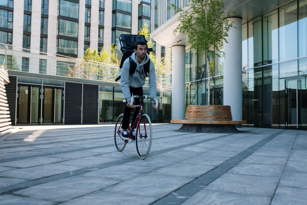 Fahrradkurier mit schwarzer Rückentasche fährt an einem Bürogebäude entlang.