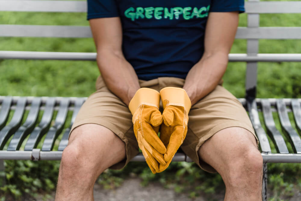 Ein Mann mit einem Greenpeace T-Shirt und gelben Arbeitshandschuhen sitzt auf einer Parkbank.