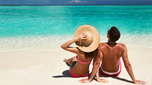 Eine Frau mit Sonnenhut und ein Mann sitzen an einem Strand und schauen in den Himmel.