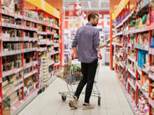 Ein junger Mann geht mit Einkaufswaagen durch den Supermarkt und schaut sich Produkte genau an.