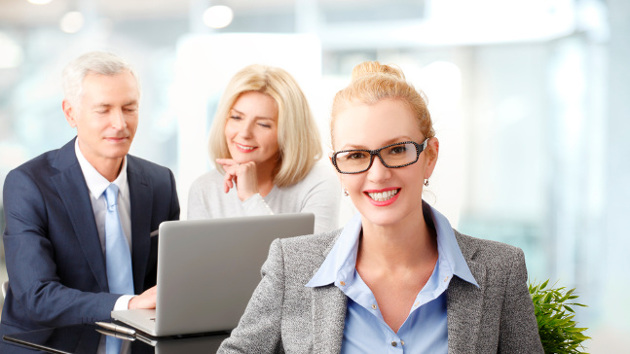 Eine Frau in Berufskleidung sitzt vor zwei am Laptop arbeitenden Personen und lächelt.