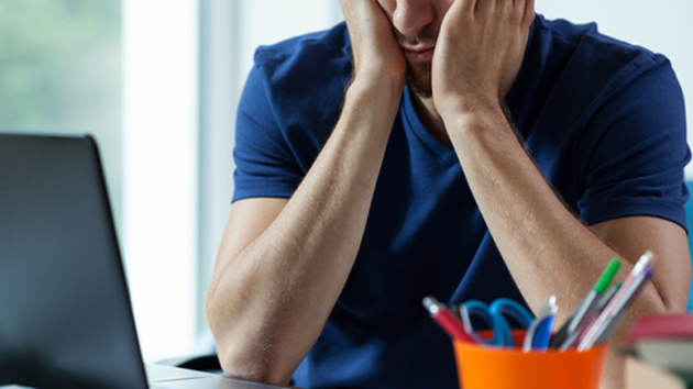 Ein Mann in blauem T-Shirt sitzt vor seinem Laptop und stützt seinen Kopf auf seine Hände.