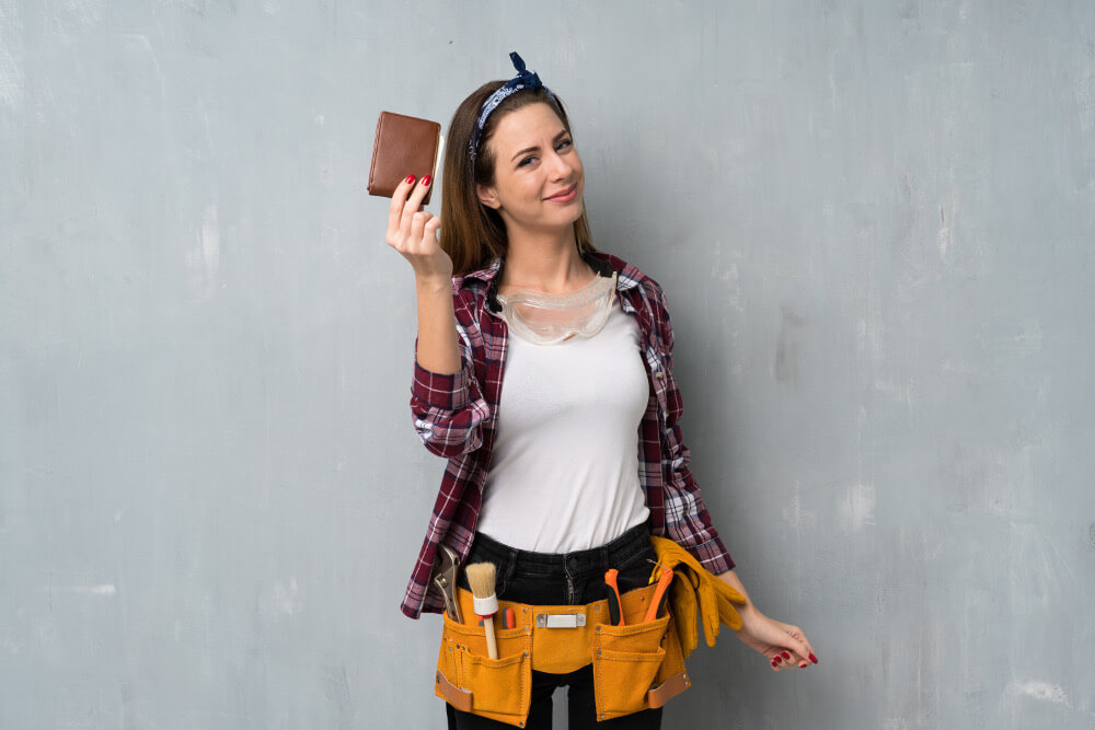 Eine junge Handwerkerin mit Werkzeuggürtel hält erfreut ein Portemonnaie in die Höhe.