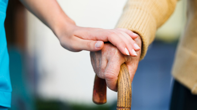 Eine Pflegekraft legt ihre Hand über die Hand einer pflegebedürftigen Person, die sich auf einen Gehstock stützt.