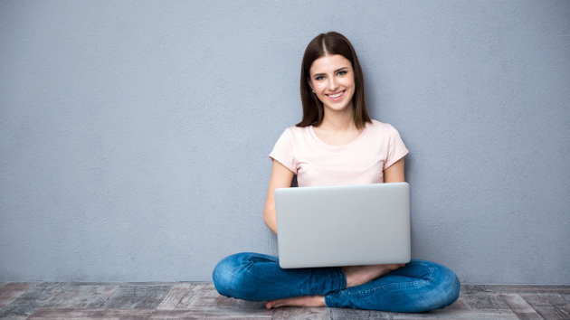 Eine lächelnde Frau sitzt mit ihrem Laptop auf dem Boden vor einer grauen Wand.