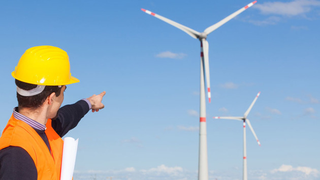 Ein Mann in Arbeitskleidung zeigt mit dem Finger auf zwei Windkraftanlagen.