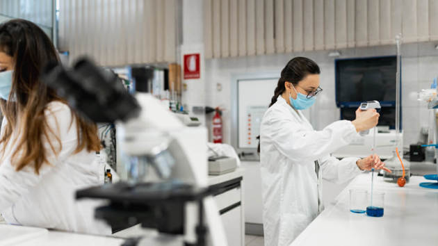 Wissenschaftlerin im weißen Kittel und mit Schutzmaske führt ein chemisches Experiment mit einer blauen Flüssigkeit in einem modernen Labor durch
