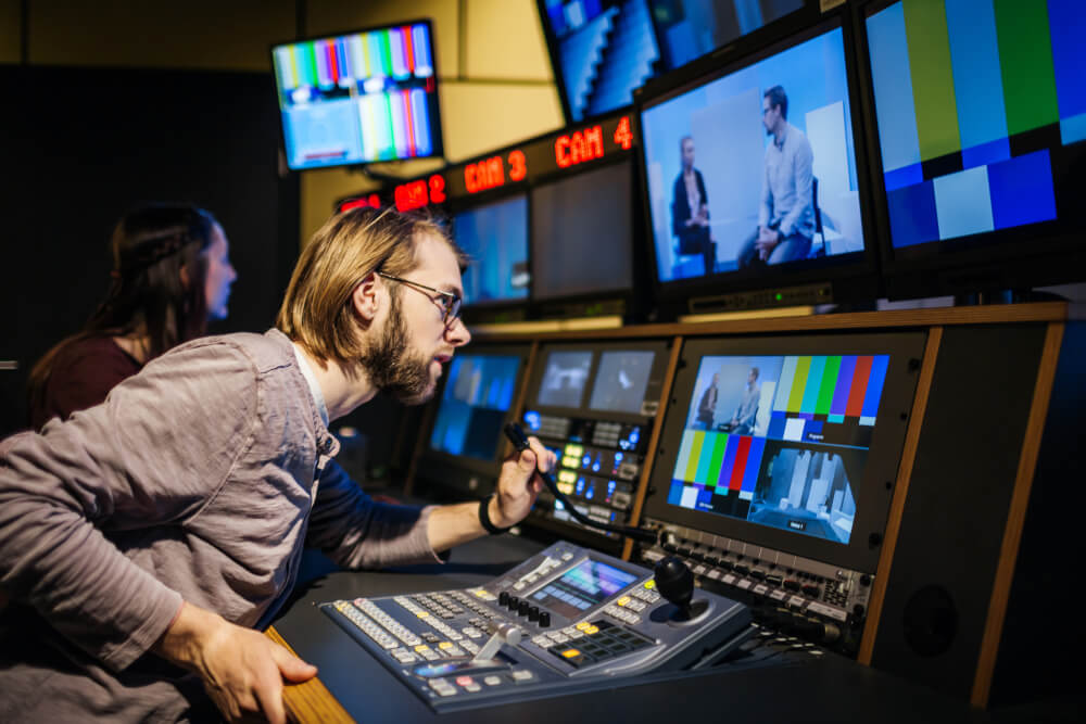 Mann gibt Teilnehmern einer Fernsehshow Anweisungen über ein Mikrofon, während er sie über einen Bildschirm beobachtet.