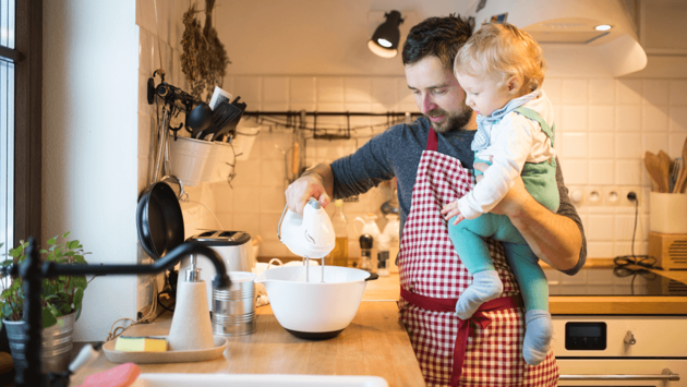 Ein Mann steht in der Küche, hält ein Kleindkind auf dem Arm und hält einen Mixer in eine Rührschüssel.