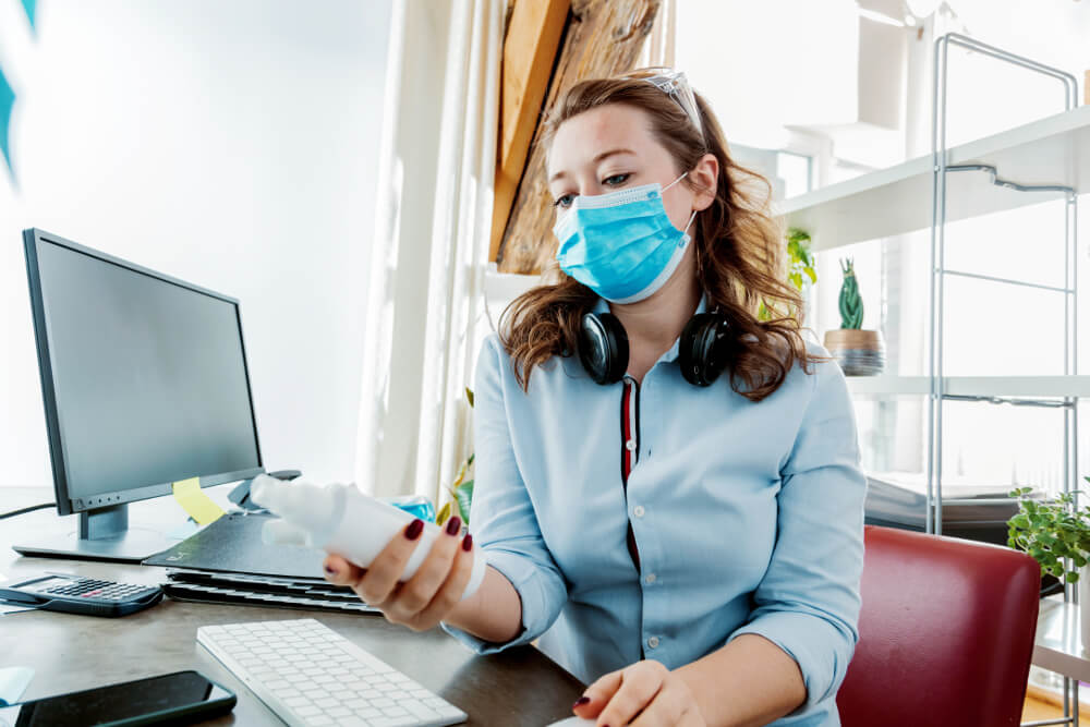 Junge Frau mit Mund-Nasen-Schutz und Desinfektionsspray an ihrem Arbeitsplatz in einem Büro.